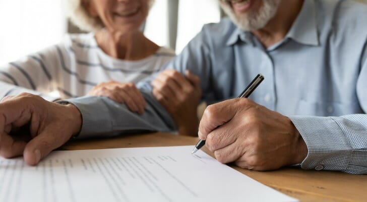 Senior man signing legal document at table with supportive woman smiling beside him.